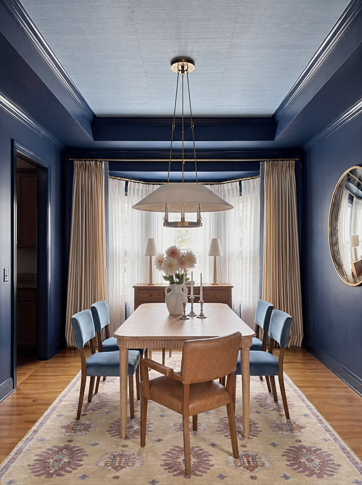 Navy dining room with a tray ceiling painted Hale Navy and sky blue grasscloth on the ceiling, a shaded chandelier over a cerused oak table with blue velvet chairs, camel striped curtains, Eleanor travertine buffet lamps and the KFD Cairene hand-knotted rug