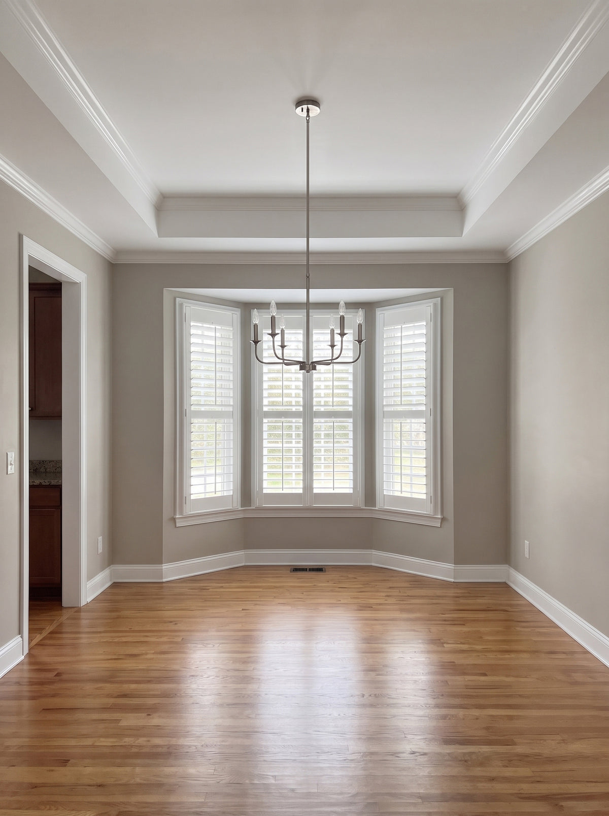 Builder-grade dining room with a tray ceiling, its soffit and ceiling painted white over greige walls, a brushed nickel chandelier, plantation shutters in a bay window and bare red oak floors