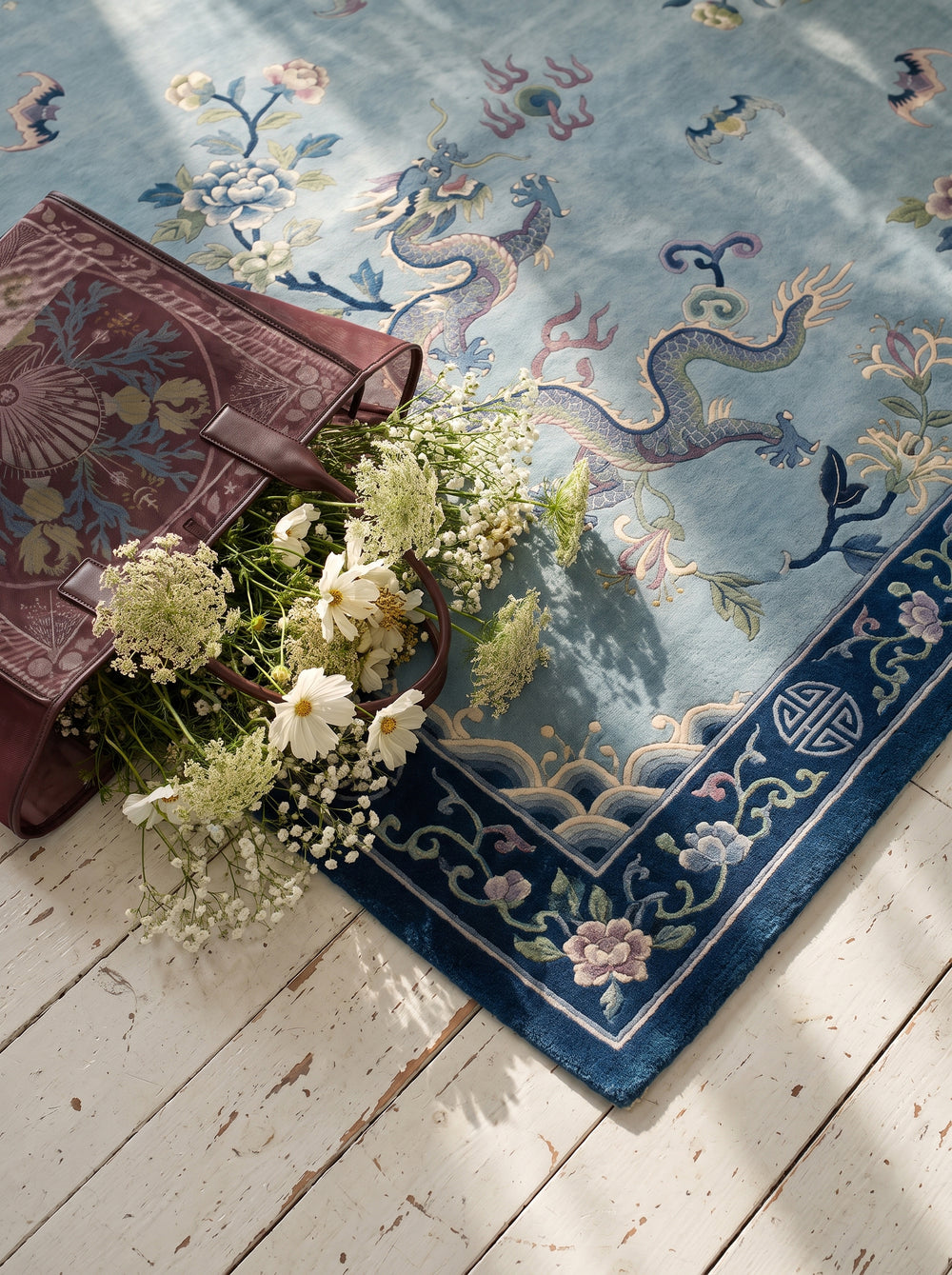 A light blue Chinese Art Deco area rug with dragons and floral borders, styled on a white wooden floor with white flowers.