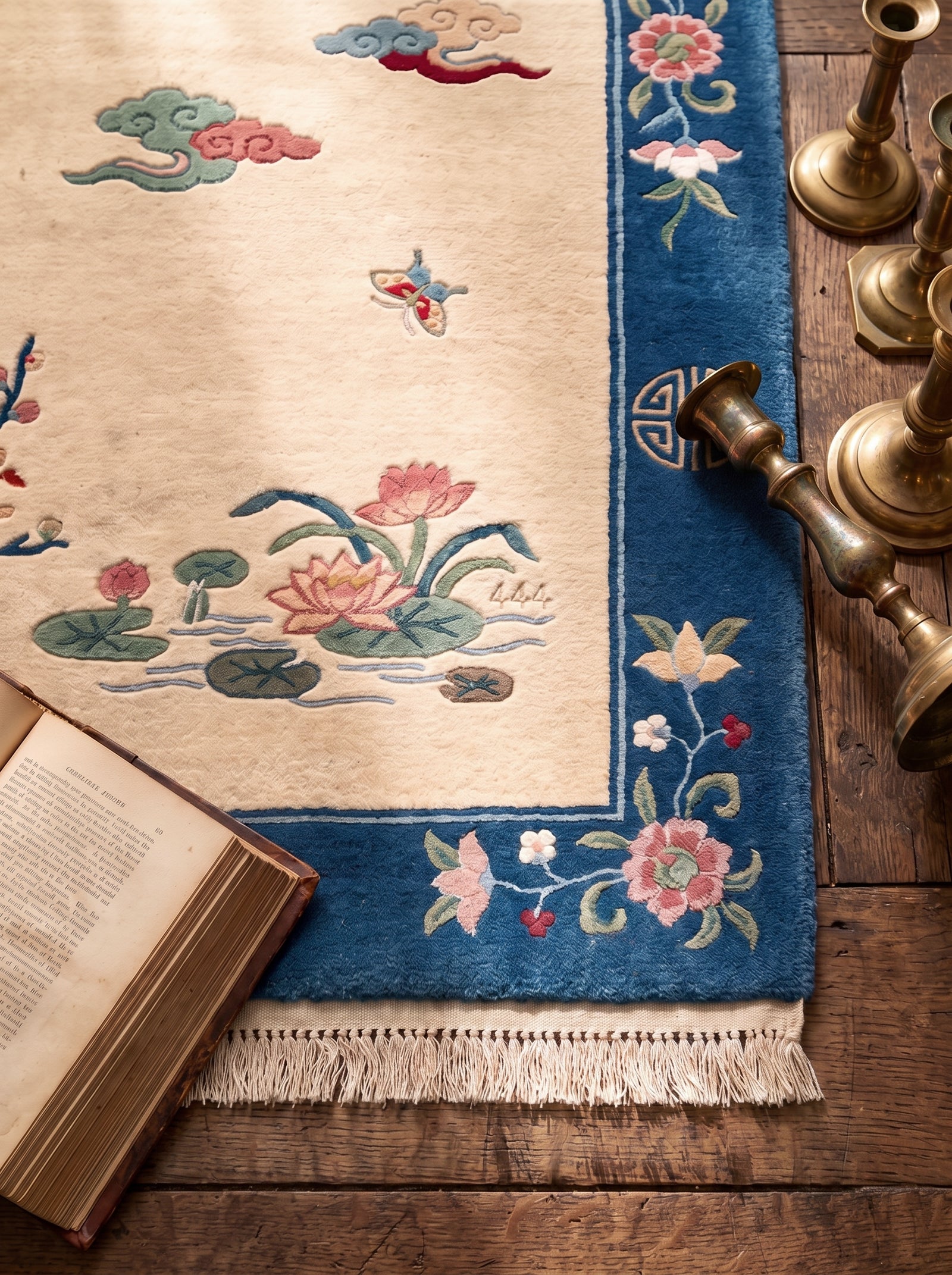 A custom Chinese Art Deco horoscope area rug with a beige field, blue border, and lotus and cloud motifs, styled on a wooden floor with an open book and brass candlesticks.